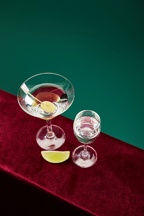 high angle view of shot glass and cocktail glass with vermouth, lime slice and whole olive on toothpick on velour surface isolated on green