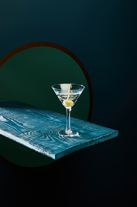 cocktail glass with vermouth and whole olive on toothpick on blue wooden surface on geometric background