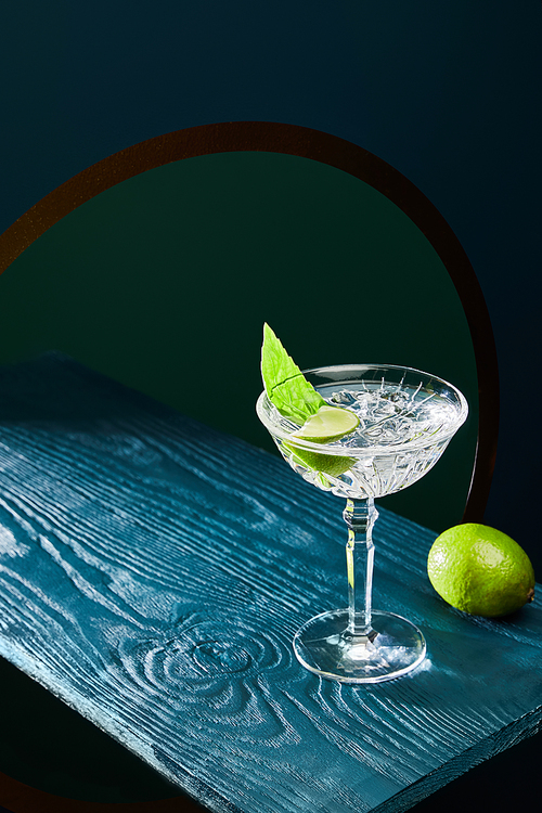 High angle view of cocktail glass with mint leaf and whole lime on blue wooden surface on geometric background with circle