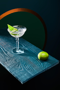 high angle view of cocktail glass with mint leaf and whole lime on blue wooden surface on geometric background with golden circle