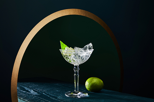 cocktail glass with mint leaf and whole lime on blue wooden surface on geometric background with golden circle