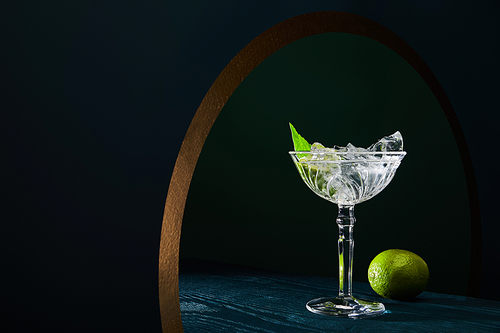 cocktail glass with ice, mint leaf and whole lime on blue wooden surface on background with golden circle