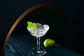 high angle view of cocktail glass with ice, mint leaf and whole lime on blue wooden surface on geometric background with golden circle