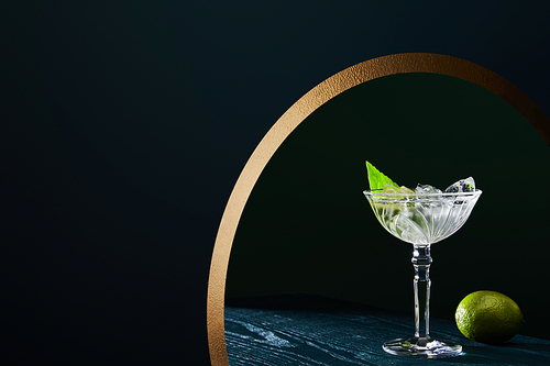 cocktail glass with ice cubes, mint leaf and whole lime on blue wooden surface on geometric background with golden circle