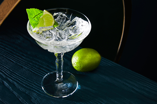 High angle view of cocktail glass with ice cubes, mint leaf and whole lime on blue wooden surface on geometric background