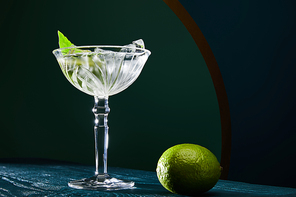 low angle view of cocktail glass with ice cubes, mint leaf and whole lime on blue wooden surface on geometric background