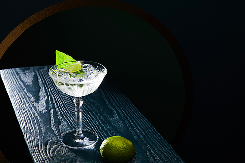 high angle view of cocktail glass with drink with ice cubes, mint leaf and whole lime on blue wooden surface on black background