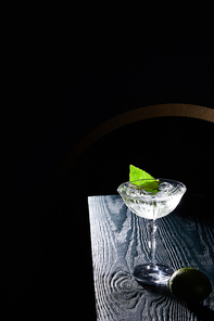 high angle view of cocktail glass with drink with ice cubes, mint leaf and whole lime on blue wooden surface on black background with geometric lines