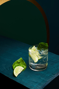 high angle view of old fashioned glass with fresh drink, mint leaf and lime slice on blue wooden surface on green and blue geometric background