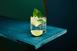 high angle view of old fashioned glass with refreshing drink, mint leaf and lime slice on blue wooden surface on green and blue geometric background
