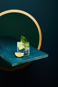 High angle view of old fashioned glass with fresh drink, mint leaf and lime slice on blue wooden surface on green and blue geometric background with golden circle
