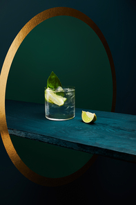 Glass with refreshing drink with ice cubes, mint leaf and lime slice on wooden surface on geometric blue and green background