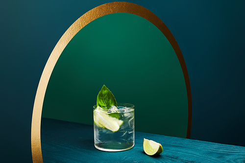 High angle view of glass with refreshing drink with lime, ice cubes and mint leaf on wooden surface on geometric blue and green background