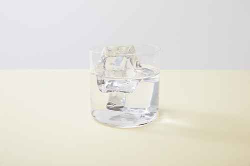 Glass with water and ice cubes on yellow surface isolated on grey