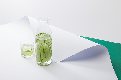 high angle view of bottle and glass with fresh drink and sliced cucumbers on white paper surface on grey and green background