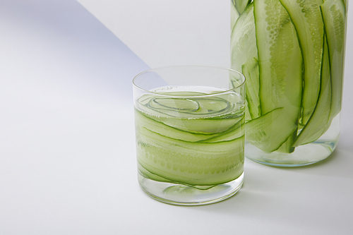 Close up view of bottle and glass with fresh drink and sliced cucumbers on grey and white background