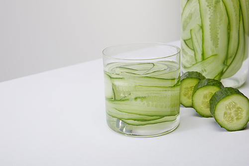Close up view of cropped bottle and glass with fresh detox drink and sliced cucumbers isolated on grey