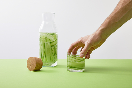 Cropped view of male hand holding glass with drink made of sliced cucumbers and bottle isolated on grey