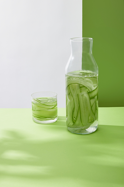 Bottle and glass with fresh drink made of sliced cucumbers on grey and green background