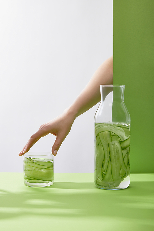 Cropped view of female hand touching glass with fresh drink made of sliced cucumbers and bottle on grey and green background