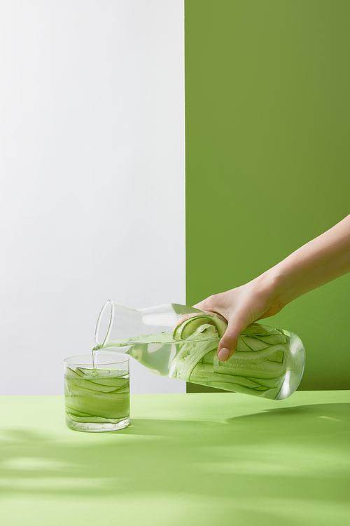 Cropped view of female hand pouring water from bottle into glass with sliced cucumbers on grey and green background