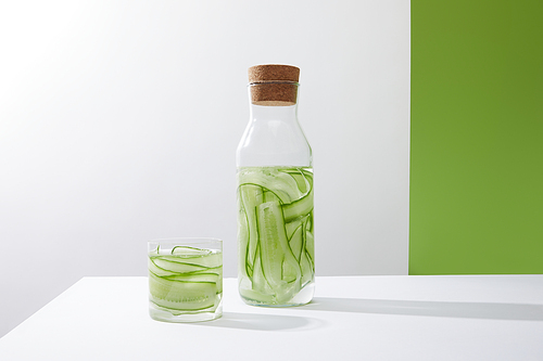 Bottle with cork and glass with drink made of sliced cucumbers on grey and green background
