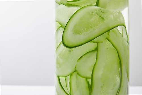 Close up view of glass bottle filled with liquid and sliced cucumbers on grey and white background