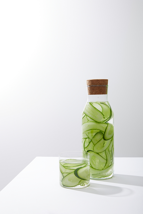 Glass and bottle with cork filled with water and sliced cucumbers on grey background