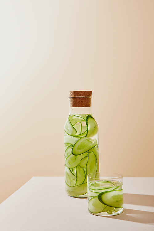 Glass and bottle with cork filled with water and sliced cucumbers on table on beige background