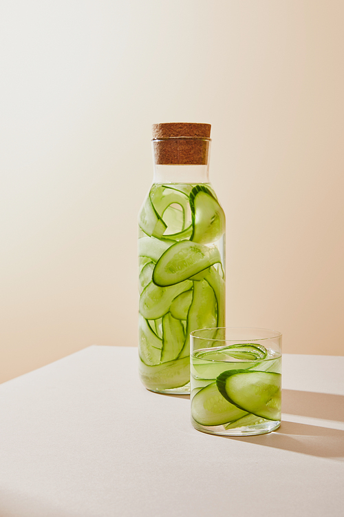 Glass and bottle with cork filled with fresh water and sliced cucumbers on beige background