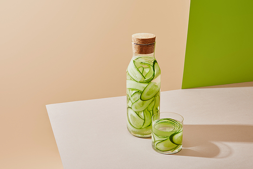 high angle view of glass and bottle with cork filled with fresh water and sliced cucumbers on table on beige and green background