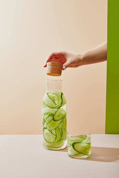Cropped view of female hand holding cork under bottle filled with water and sliced cucumbers on table on beige and green background