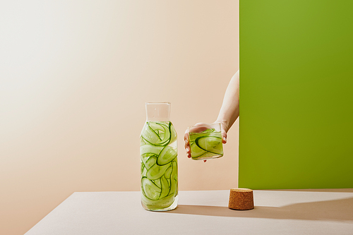 Cropped view of female hand holding glass filled with water and sliced cucumbers on beige and green background