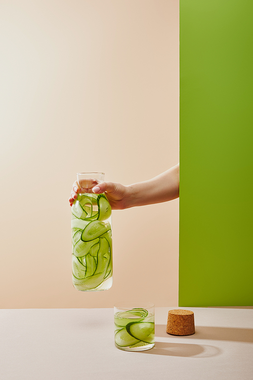 Cropped view of female hand holding bottle filled with water and sliced cucumbers on beige and green background