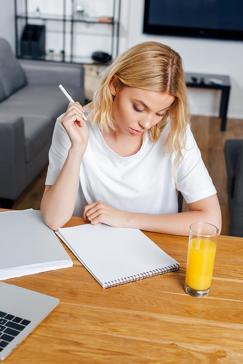 Blonde girl holding pen near notebook, laptop and glass of orange juice at home