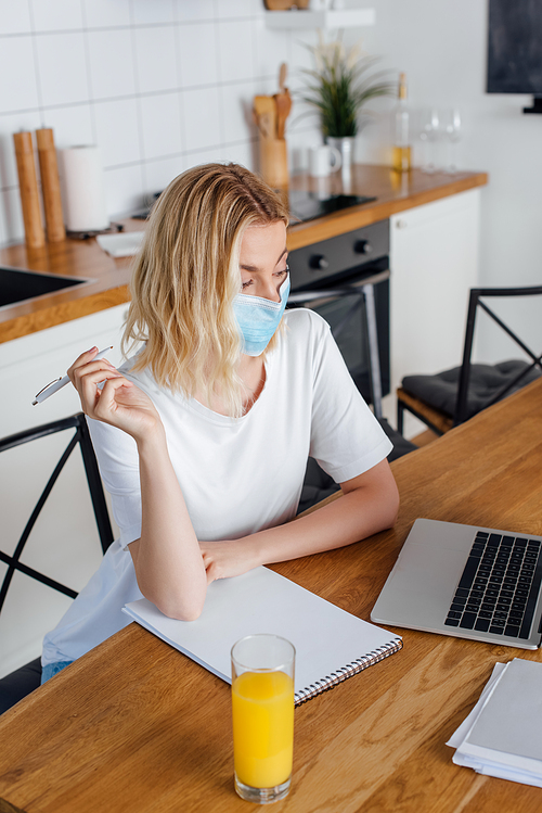 Freelancer in medical mask holding pen near orange juice, laptop and notebook on table