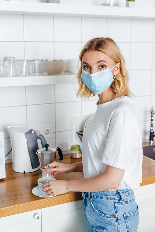 Side view of woman in medical mask  while mixing coffee in kitchen