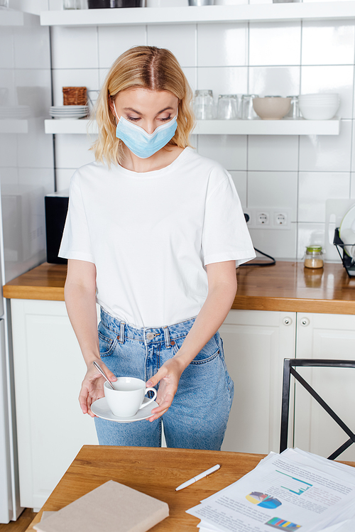 young woman in medical mask holding saucer and cup of coffee