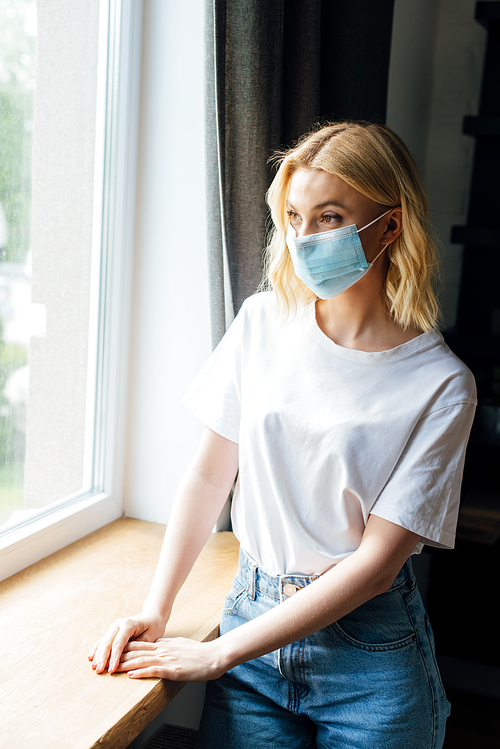 young woman in medical mask standing near window at home