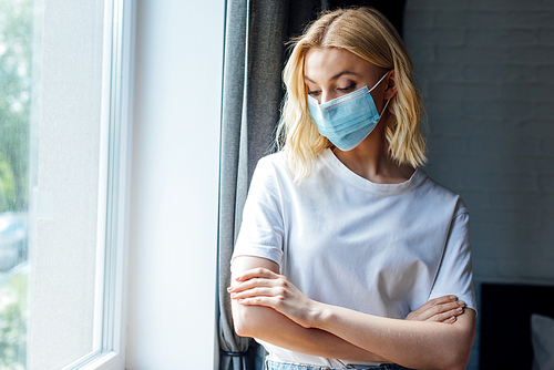 young woman in medical mask with crossed arms standing near window at home