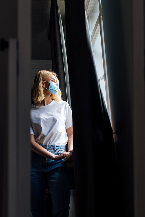 selective focus of young woman in medical mask standing near window at home