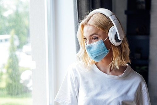 young woman in medical mask and wireless headphones standing near window at home