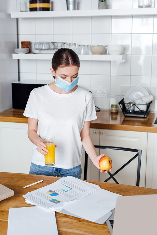 selective focus of young freelancer in medical mask holding glass of orange juice and apple near laptop