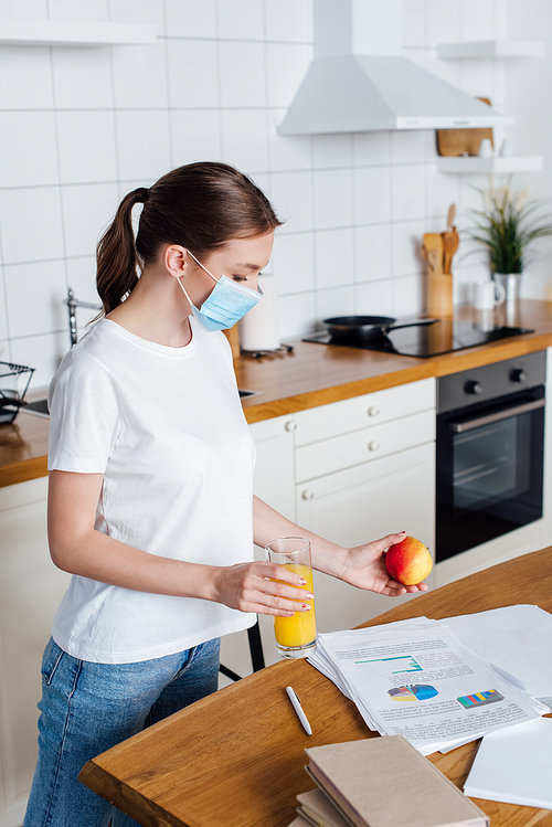 young woman in medical mask holding glass of orange juice and apple near table with charts and graphs