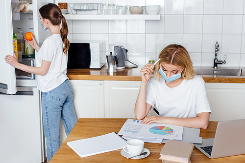 woman holding apple near fridge and sister in medical mask working from home