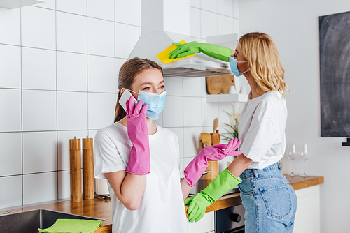 selective focus of woman in medical mask talking on smartphone near sister cleaning kitchen