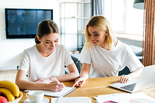 cheerful woman looking at sister writing in notebook neat laptop, online study concept