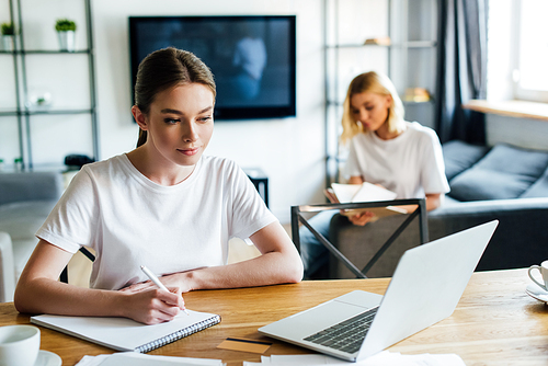 selective focus of woman looking at laptop while writing in notebook near sister, online study concept