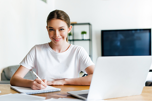 selective focus of happy woman holding pen near notebook and laptop