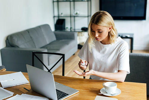 young woman holding bottle with hand sanitizer near laptop and credit card on table